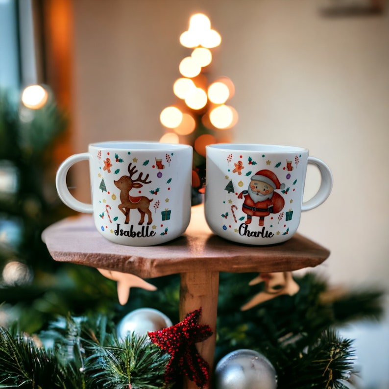以下が含まれることがあります： クリスマスのデザインが施された白いセラミック製のマグカップが2つ。1つのマグカップには「Isabelle」という名前のトナカイが描かれ、もう1つのマグカップには「Charlie」という名前のサンタクロースが描かれています。