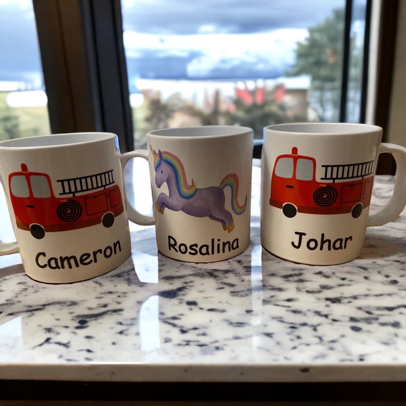 Op de afbeelding: Drie witte keramische mokken met gepersonaliseerde namen. De mokken hebben een rode brandweerwagen, een paars eenhoorn en een rode brandweerwagen. De mokken staan op een wit aanrecht.