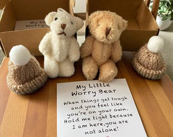 May include: Two small teddy bears, one cream and one tan, sit next to a card that reads "My Little Worry Bear." Each bear wears a knitted hat with a white pom-pom. The bears are displayed with two small cardboard boxes.