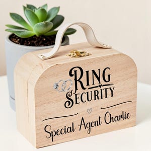 May include: A light-coloured wooden box with a white handle and a gold latch. The front features black text that reads "RING SECURITY" and "Special Agent Charlie," along with a graphic of two interlocking rings and a small heart icon.