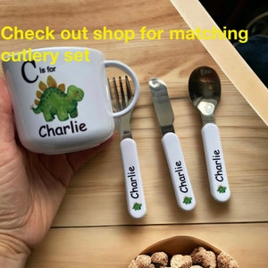 Op de afbeelding: Een witte keramische mok met een groene dinosaurus illustratie en de tekst "C is voor Charlie". De mok maakt deel uit van een bijpassend bestekset met een vork, mes en lepel, allemaal met witte handvatten en de naam "Charlie" erop gedrukt.