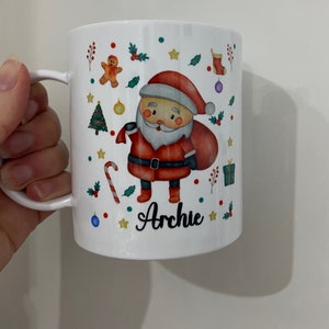 以下が含まれることがあります： サンタクロース、キャンディーケーン、クリスマスツリー、その他のクリスマスの要素が描かれた白いセラミックマグカップ。マグカップには「アーチー」という名前が書かれています。