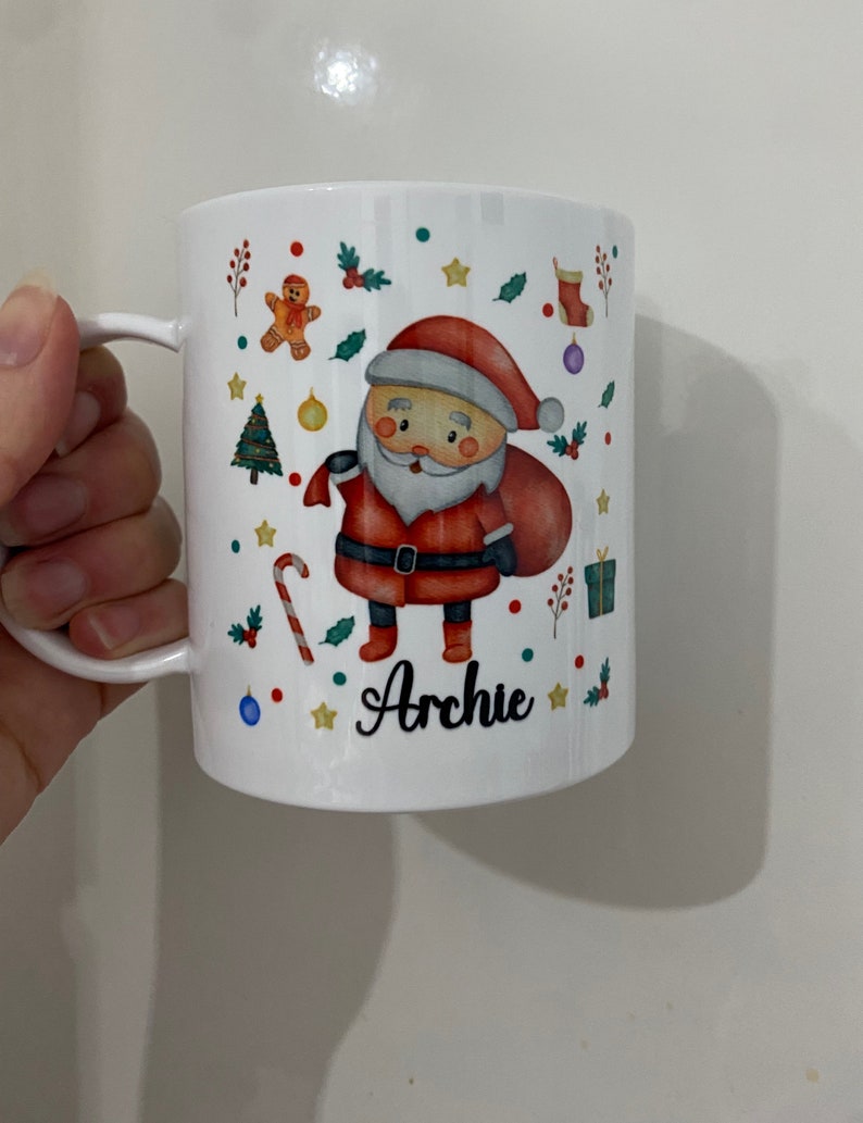 以下が含まれることがあります： サンタクロース、キャンディーケーン、クリスマスツリー、その他のクリスマスの要素が描かれた白いセラミックマグカップ。マグカップには「アーチー」という名前が書かれています。