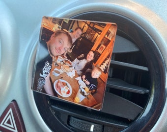 Op de afbeelding: Een vierkante, heldere fotomagneet met een gezin aan een restauranttafel. De magneet is bevestigd aan een ventilatierooster van een auto. De foto toont een man, twee vrouwen en een kind aan een tafel met borden eten.