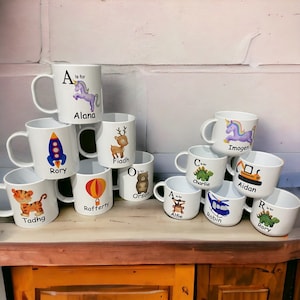 Op de afbeelding: Een verzameling witte keramische mokken met kleurrijke illustraties en namen op elke mok. De mokken tonen een verscheidenheid aan dieren, waaronder een eenhoorn, een hert, een beer, een aap, een dinosaurus, een raket, een luchtballon, een tijger, een helikopter en een bouwvoertuig. De mokken zijn gestapeld en gerangschikt op een houten oppervlak.