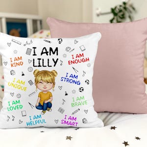 May include: White decorative cushion featuring a cartoon girl illustration and the text "I AM LILLY". The cushion also has the phrases "I AM KIND", "I AM ENOUGH", "I AM UNIQUE", "I AM STRONG", "I AM LOVED", "I AM BRAVE", "I AM HELPFUL", and "I AM SMART". A solid pink cushion is behind it.