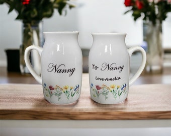 May include: Two white ceramic mugs with floral designs and black text. The mug on the left says "Nanny". The mug on the right says "To Nanny Love Amelia".