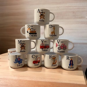 Op de afbeelding: Een set van 10 witte keramische mokken met kleurrijke illustraties en letters. Elke mok heeft een andere letter van het alfabet en een bijbehorend dier of voertuig. De mokken zijn in een piramidevorm gestapeld.