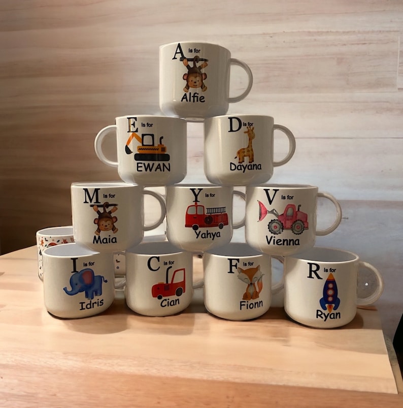 Op de afbeelding: Een set van 10 witte keramische mokken met kleurrijke illustraties en letters. Elke mok heeft een andere letter van het alfabet en een bijbehorend dier of voertuig. De mokken zijn in een piramidevorm gestapeld.