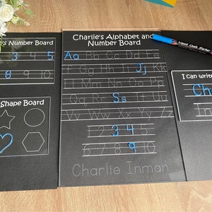 May include: Three black dry-erase boards with white outlines. The first board has the numbers 1-10 written on it. The second board has the alphabet and numbers 1-10 written on it. The third board has the words "I can write my name" and the name "Charlie Inman" written on it. A blue liquid chalk marker is lying on the boards.