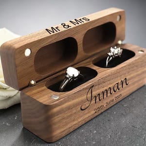 May include: A wooden ring box with two compartments, one holding a diamond ring. The box is engraved with "Mr & Mrs" on the lid and "Inman 28th June 2025" on the front. The box is open, revealing the rings.