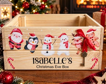 May include: A light-colored wooden crate featuring a watercolor illustration of Santa, a penguin, a snowman, a polar bear, a reindeer, and a gingerbread man. The text reads Isabelle's Christmas Eve Box, accented by a red ribbon and a metal key.