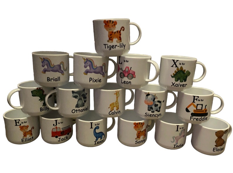 Op de afbeelding: Een verzameling van 14 witte keramische mokken met kleurrijke dier- en letterontwerpen. Elke mok heeft een andere naam gedrukt op de bodem, zoals "Elliot", "Jack" en "Indie".
