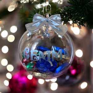 May include: Clear glass Christmas bauble with a silver bow and the name "GEMMA" written on it. The bauble is filled with small, colourful sweets.