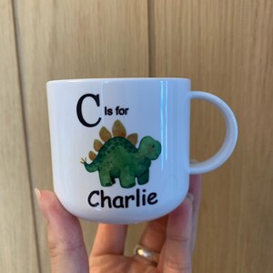 Op de afbeelding: Een witte keramische mok met een groene dinosaurus illustratie en de tekst "C is voor Charlie".