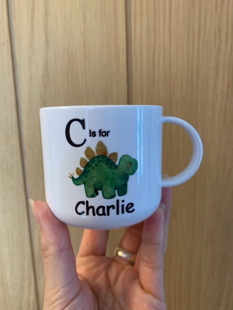 Op de afbeelding: Een witte keramische mok met een groene dinosaurus illustratie en de tekst "C is voor Charlie".