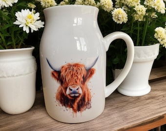 May include: White ceramic mug with a brown and white Highland cow illustration. The mug has a large handle and a rounded shape.