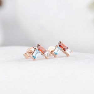 May include: A pair of gold earrings with a cluster of red, blue, and clear gemstones. The earrings are set in a simple, minimalist style.