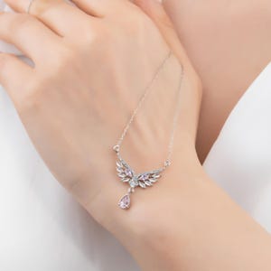 Peut inclure: Un collier argent&eacute; avec un pendentif en forme d'ailes. Le pendentif pr&eacute;sente un groupe central de pierres roses, entour&eacute; de cristaux transparents. Une pierre rose en forme de goutte pend en dessous. Le collier est pr&eacute;sent&eacute; sur une main.