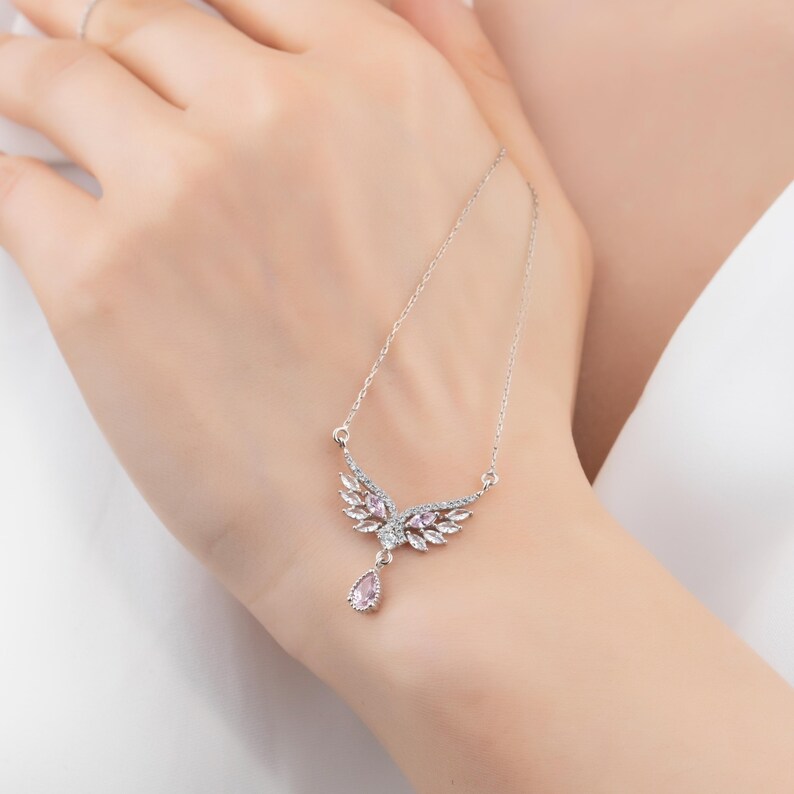 Peut inclure: Un collier argent&eacute; avec un pendentif en forme d'ailes. Le pendentif pr&eacute;sente un groupe central de pierres roses, entour&eacute; de cristaux transparents. Une pierre rose en forme de goutte pend en dessous. Le collier est pr&eacute;sent&eacute; sur une main.