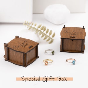 Peut inclure: Trois bagues avec des pierres pr&eacute;cieuses de couleurs diff&eacute;rentes sont pr&eacute;sent&eacute;es &agrave; c&ocirc;t&eacute; de bo&icirc;tes &agrave; bijoux en bois. Les bagues sont en or, argent et or rose. Le texte "Special Gift Box" est en bas.