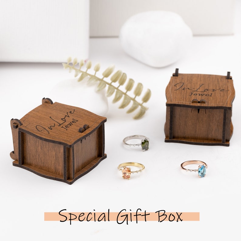 Peut inclure: Trois bagues avec des pierres pr&eacute;cieuses de couleurs diff&eacute;rentes sont pr&eacute;sent&eacute;es &agrave; c&ocirc;t&eacute; de bo&icirc;tes &agrave; bijoux en bois. Les bagues sont en or, argent et or rose. Le texte "Special Gift Box" est en bas.