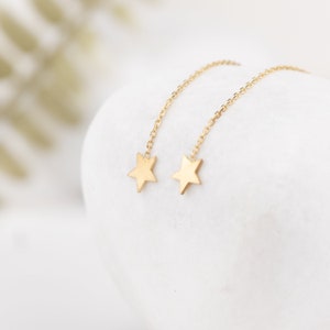 May include: A pair of gold-colored star-shaped earrings. Each earring features a small star pendant suspended from a delicate chain. The earrings are displayed against a white background, creating a minimalist aesthetic.