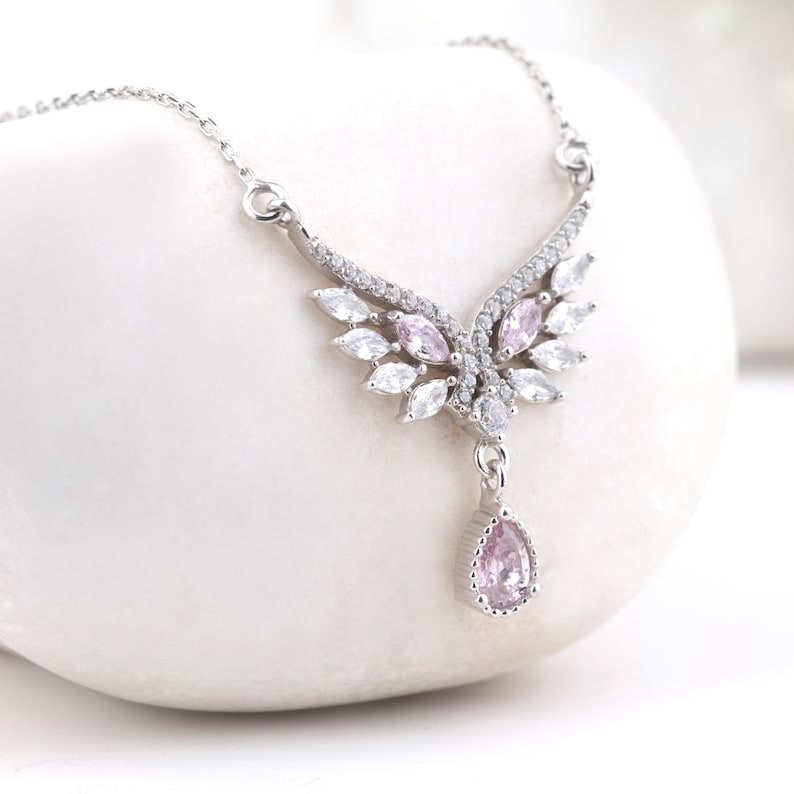Peut inclure: Un collier argent&eacute; avec un pendentif en forme d'aile. Le collier est orn&eacute; de pierres pr&eacute;cieuses blanches et roses et d'un pendentif rose en forme de goutte. Le collier est pr&eacute;sent&eacute; sur une surface blanche.