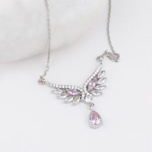 Peut inclure: Un collier argent&eacute; avec un pendentif en forme d'aile orn&eacute; de pierres pr&eacute;cieuses claires et roses. Le pendentif pr&eacute;sente un groupe de pierres roses et une pierre rose en forme de goutte. Le collier est sur fond blanc.