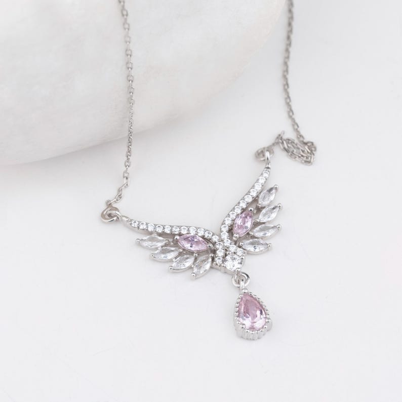 Peut inclure: Un collier argent&eacute; avec un pendentif en forme d'aile orn&eacute; de pierres pr&eacute;cieuses claires et roses. Le pendentif pr&eacute;sente un groupe de pierres roses et une pierre rose en forme de goutte. Le collier est sur fond blanc.