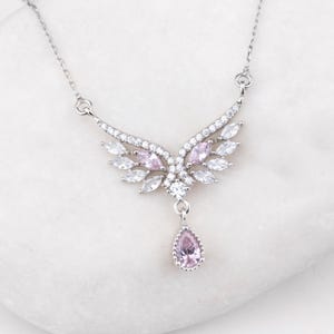 Peut inclure: Un collier argent&eacute; avec un pendentif en forme d'ailes stylis&eacute;es. Les ailes sont orn&eacute;es de pierres pr&eacute;cieuses claires et roses. Une pierre rose en forme de goutte pend du centre. Le collier est pr&eacute;sent&eacute; sur une surface blanche.