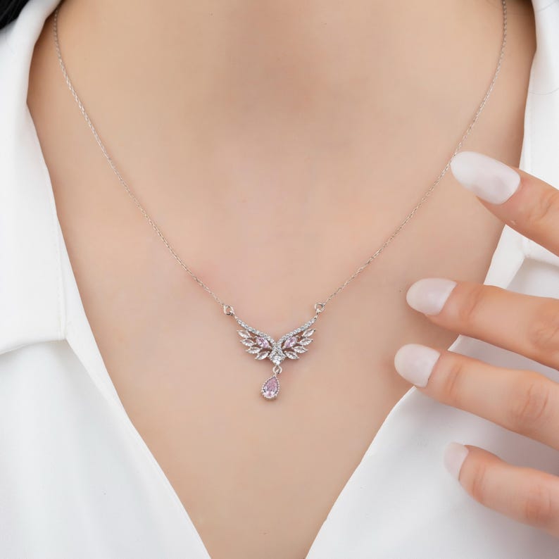 Peut inclure: Un collier argent&eacute; avec un pendentif en forme d'ailes orn&eacute; de petites pierres pr&eacute;cieuses claires et roses. Une pierre pr&eacute;cieuse rose en forme de goutte pend du centre. Le collier est port&eacute; sur une chemise blanche.