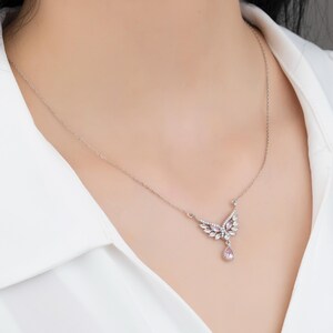 Peut inclure: Un collier argent&eacute; avec un pendentif en forme d'ailes orn&eacute; de pierres pr&eacute;cieuses claires et roses. Le collier est port&eacute; sur une chemise blanche, mettant en valeur le design d&eacute;licat et l'&eacute;clat des bijoux.