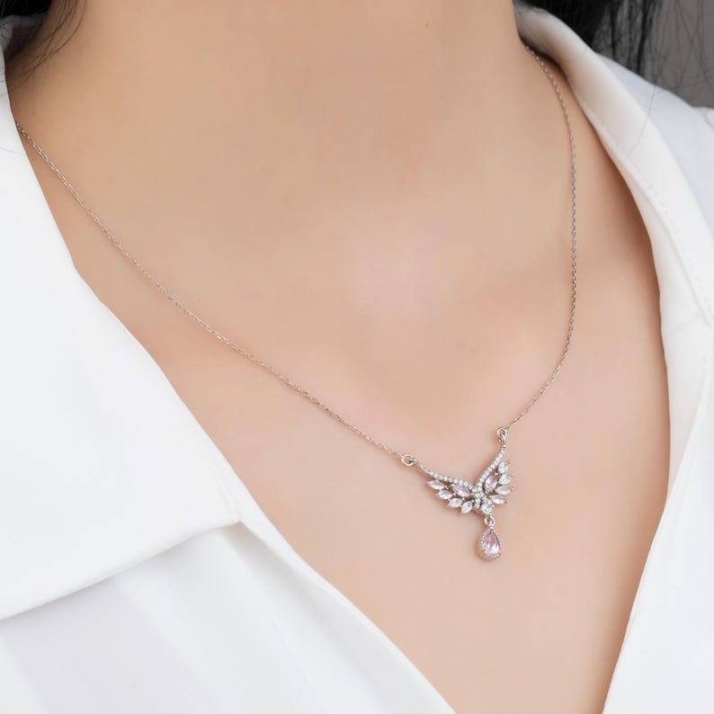 Peut inclure: Un collier argent&eacute; avec un pendentif en forme d'ailes orn&eacute; de pierres pr&eacute;cieuses claires et roses. Le collier est port&eacute; sur une chemise blanche, mettant en valeur le design d&eacute;licat et l'&eacute;clat des bijoux.