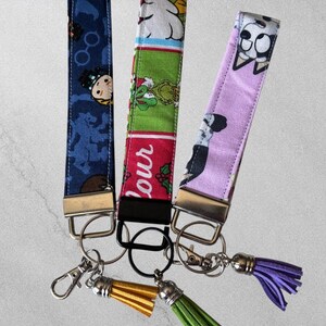 May include: Three fabric wristlet keychains with metal hardware and attached tassels. The left is dark blue with character prints, the center is green and red with text, and the right is light purple with a cartoon dog print. Each has a lobster clasp.