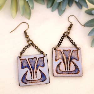 Könnte beinhalten: Ein Paar quadratische Holzohrringe mit hellblauem Hintergrund und blauem Blumendesign. Die Ohrringe sind mit einer Bronzekette und einem Haken verbunden. Das Design ist braun umrandet.