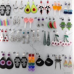 May include: A collection of various novelty earrings, including coffin-shaped designs with the text "Thick Thighs and Spooky Vibes", bat, avocado, and eyeball designs. Other earrings feature lollipops, cherries, and skeleton hands.