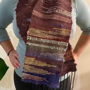 May include: A woven scarf with a colorful, abstract pattern in shades of brown, purple, yellow, and blue. The scarf has black fringe on the ends.