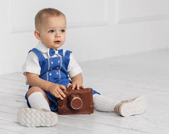 May include: A young child wearing a blue and white outfit with a bow tie holds a brown camera. The child is sitting on a white wooden floor.