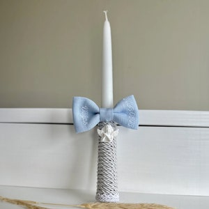 May include: A white candle decorated with a light blue bow tie with embroidered text. The candle base is wrapped in grey rope and has a small angel figurine. The bow tie has the name "Vera" and the date "25.03.03" embroidered on it.