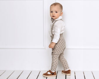 May include: A young child wearing a white button-down shirt with a bow tie and brown polka dot overalls. The child is standing on a white wooden floor and looking at the camera.