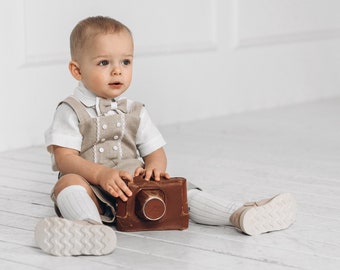 Puede incluir: Un niño pequeño con un atuendo de lino beige y una pajarita blanca se sienta en un suelo de madera blanco sosteniendo una cámara vintage marrón.