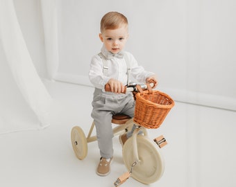 Puede incluir: Un niño pequeño con una camisa blanca y pantalones grises está sentado en un triciclo de madera con una cesta de mimbre. El triciclo tiene un asiento de cuero marrón y ruedas de color crema.