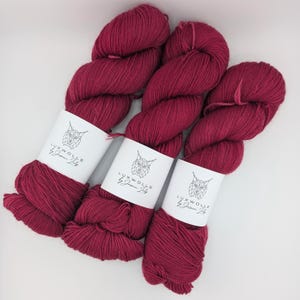 Può includere: Tre gomitoli di filato rosso scuro con etichette bianche che recitano "LUXWOLLE by Jasmin Lily"