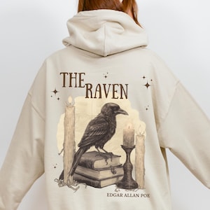 Puede incluir: Sudadera con capucha beige con un diseño de un cuervo posado sobre libros, con velas y el texto "THE RAVEN" y "EDGAR ALLAN POE." El diseño es de estilo vintage, con una ilustración detallada del ave y otros elementos.