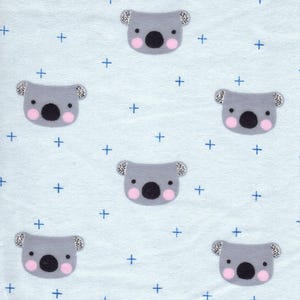 May include: Light blue fabric with a repeating pattern of gray koala faces with pink cheeks and black noses, interspersed with blue plus signs. The fabric has a ruler along the bottom edge with the text "Made in Germany".