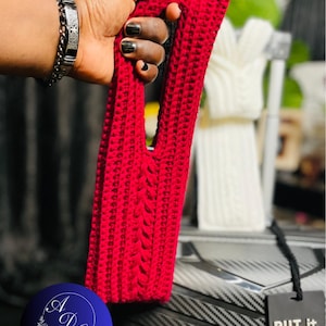 May include: A bright red crochet bag with a rectangular shape and a handle. The bag has a cable knit design on the front. The bag is held by a hand with black nail polish. A tag reads "PUT IT BACK, it's MINE!"