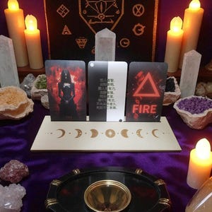 Puede incluir: Una exhibición espiritual con cartas del tarot, cristales y velas. Las cartas se muestran en un soporte de madera con diseños de fases lunares. Las cartas tienen imágenes y texto, incluida la palabra "FIRE". La escena está sobre una tela morada.