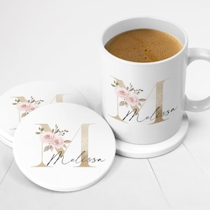 May include: White ceramic mug with a gold letter "M" and floral design. The mug says "Melissa" and is sitting on a white coaster. There are two additional white coasters with the same design.