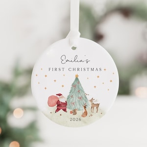K&ouml;nnte beinhalten: Ein runder, wei&szlig;er Keramik-Weihnachtsanh&auml;nger an einem wei&szlig;en Band. Das Design zeigt eine Aquarellillustration vom Weihnachtsmann, einem Weihnachtsbaum, einem Reh und einem Eichh&ouml;rnchen sowie den Text "Emilia's FIRST CHRISTMAS" und "2026".